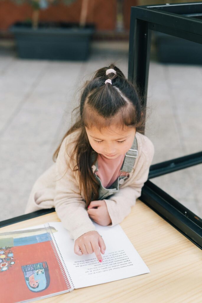 A young girl reading a book outside, focusing on the text with curiosity and concentration.