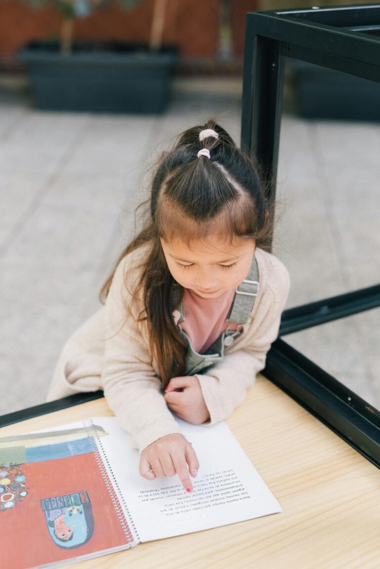 A young girl reading a book outside, focusing on the text with curiosity and concentration.
