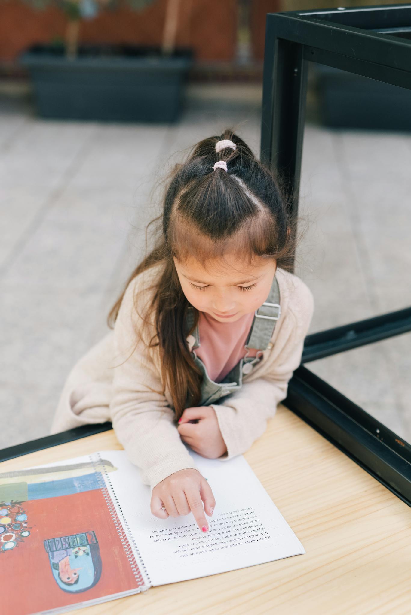A young girl reading a book outside, focusing on the text with curiosity and concentration.